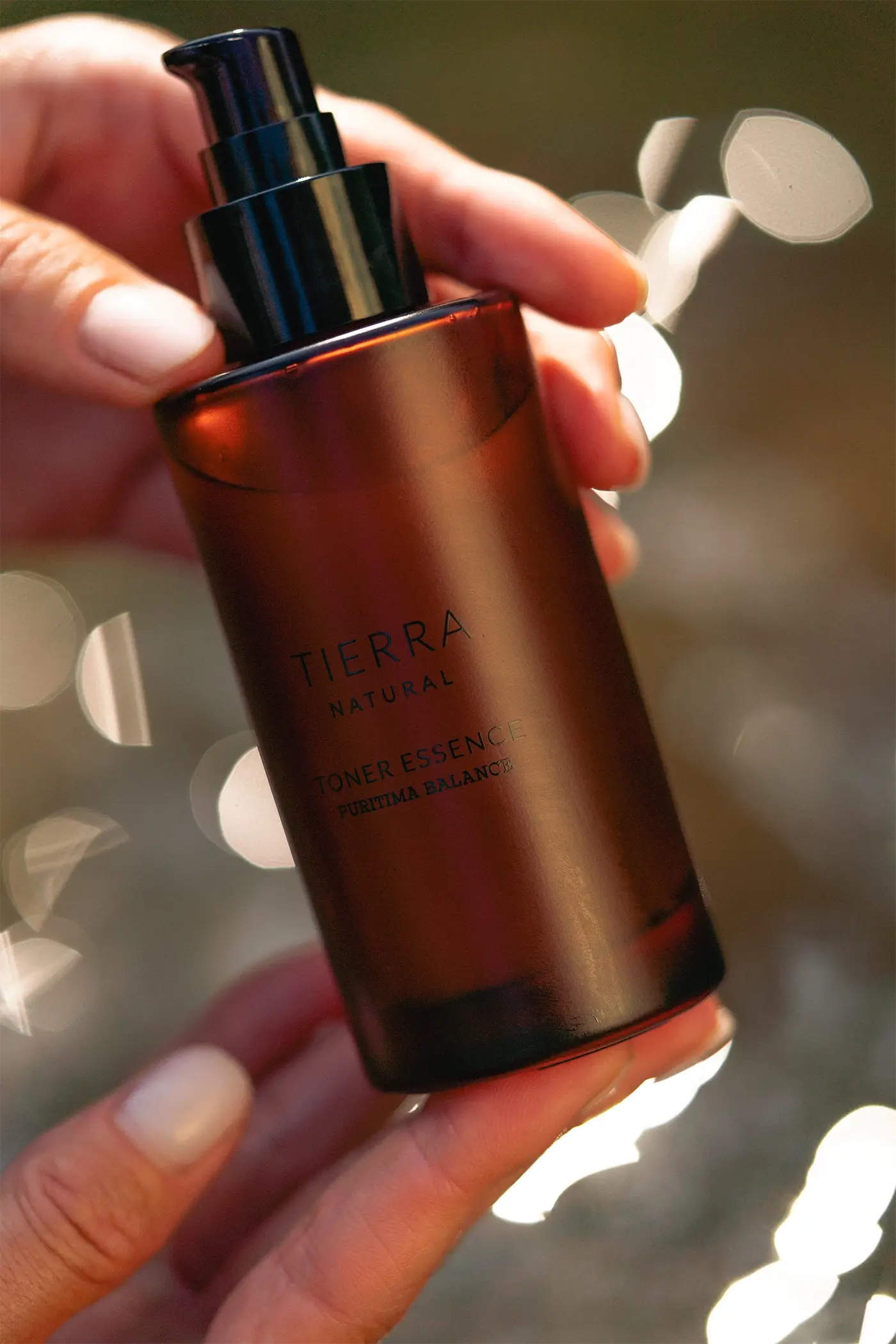 Dłonie trzymające Tierra Natural Toner Essence Purima Balance w brązowej butelce z pompką, naturalny tonik do twarzy z roślinnymi składnikami
